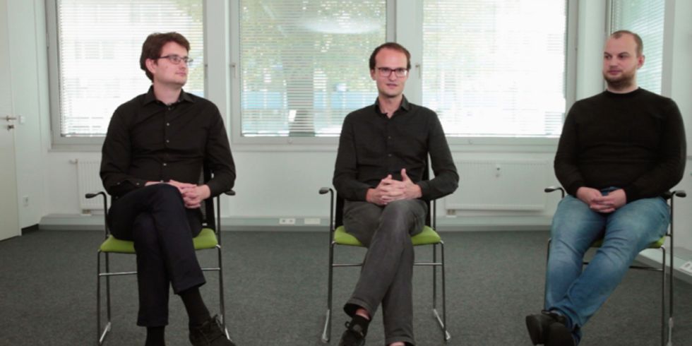 Drei Männer sitzen auf Stühlen in einem leeren Raum, einer spricht