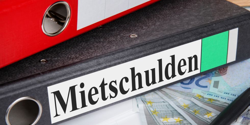 Ein Aktenordner mit der Aufschrift 'Mietschulden' liegt auf einem Schreibtisch. Auf ihm liegt ein weiterer Ordner, daneben ein Stift und mehrere Geldscheine.