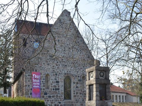 Dorfkirche Alt-Buckow Gesamtansicht von hinten: schlichte Kirche mit Sichtmauerwerk, eckigem Kirchturm und hohem Satteldach-Kirchenschiff mit Kriegsdenkmal und Büschen im Vordergrund