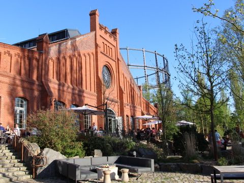 Ein rotes Backsteingebäude, vor dem Tische unter Sonnenschirmen stehen. Im Hintergrund das Gerüst eines Gasometers.