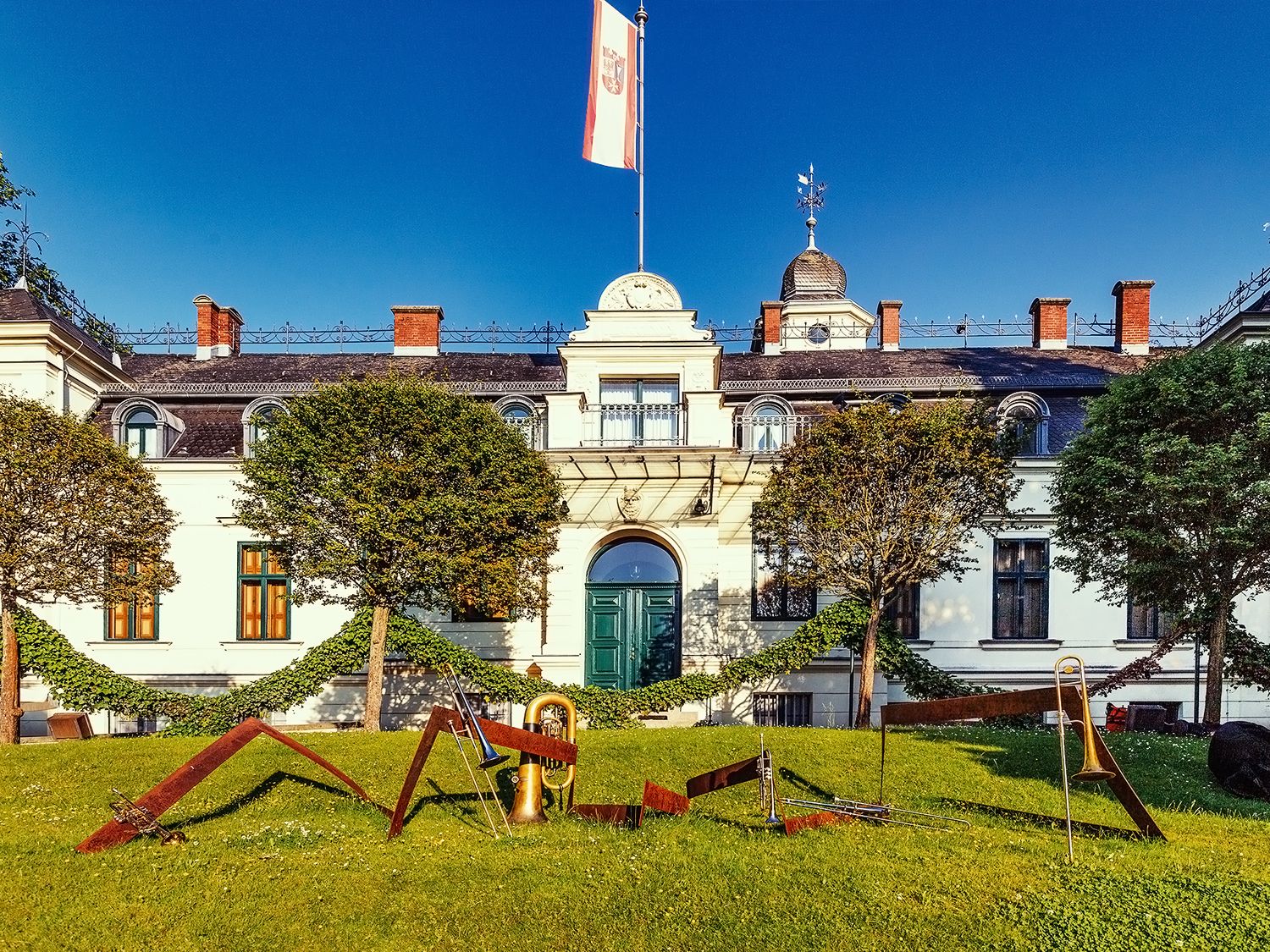 Das Schloss Britz auf einer grünen Wiese. Ein weißer Gründerzeitbau mit einem schwarzen Dach und kleinen Türmchen. Darauf weht die tor, weiß, rote Fahne Neuköllns mit dem Stadtwappen. Auf der Wiese vor dem Schloss sind in einer rostigen Kunstinstalation Blechblasinstrumente aufgebaut.