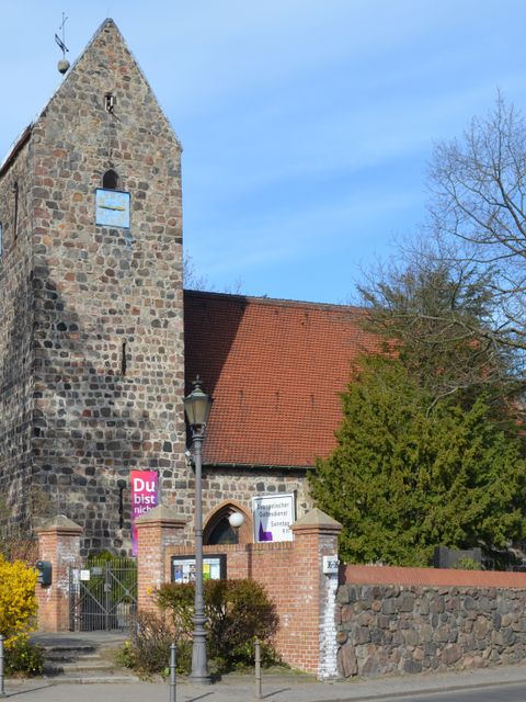Dorfkirche Alt-Buckow Gesamtansicht von der Straße: schlichte Kirche mit Sichtmauerwerk, eckigem Kirchturm und Satteldach-Kirchenschiff, eingemauert mit Bäumen und Büschen im Vordergrund