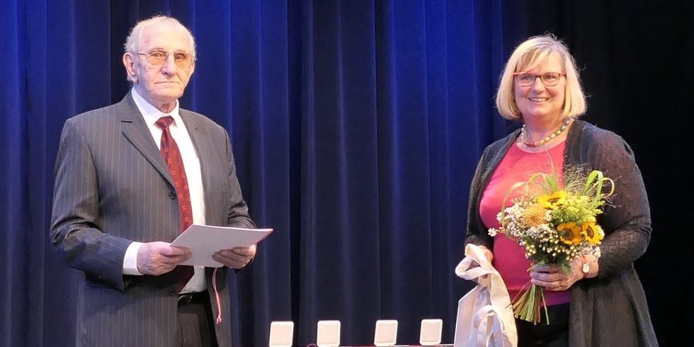 Eine Frau und ein Mann stehen nebeneinander. Der Mann hält eine Urkunde und die Frau einen Blumenstrauß und einen Beutel.