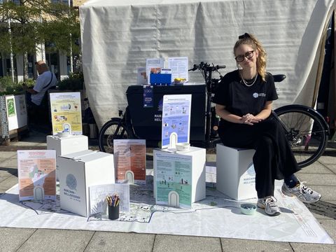 Aufbau des Infostandes des Raums für Beteiligung: Auf einer Bodenkarte sind verschiedene Sitzhocker sowie Erklärplakate zu Beteiligungsmöglichkeiten im Bezirk platziert. Im Hintergrund ist das Lastenrad zu sehen.