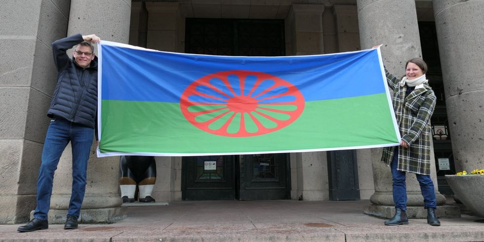 Ein Mann und eine Frau halten eine blau-grüne Flagge mit einem roten Rad hoch.