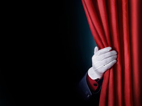 Das Bild zeigt einen Bühnenvorhang der von einer Hand beiseite geschoben wird