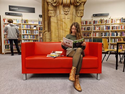 Frau mit Zeitschrift auf einem roten Sofa vor Bibliotheksregalen