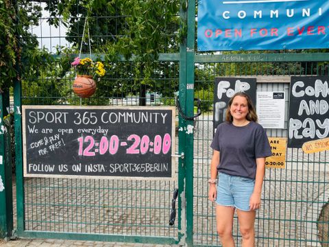 Auf dem Bild sieht man den Eingang eines Sportplatzes mit grünen Gittern und mehreren Schildern, darunter „SPORT 365 COMMUNITY“ und „OPEN FOR EVERYONE“. Die Öffnungszeiten sind von 12:00 bis 20:00 Uhr, und es wird dazu eingeladen, kostenlos mitzuspielen. Eine junge Frau steht rechts vor dem Tor, trägt ein dunkles T-Shirt und Jeansshorts. Das Gelände ist von Bäumen umgeben und wirkt einladend und offen für Besucher.