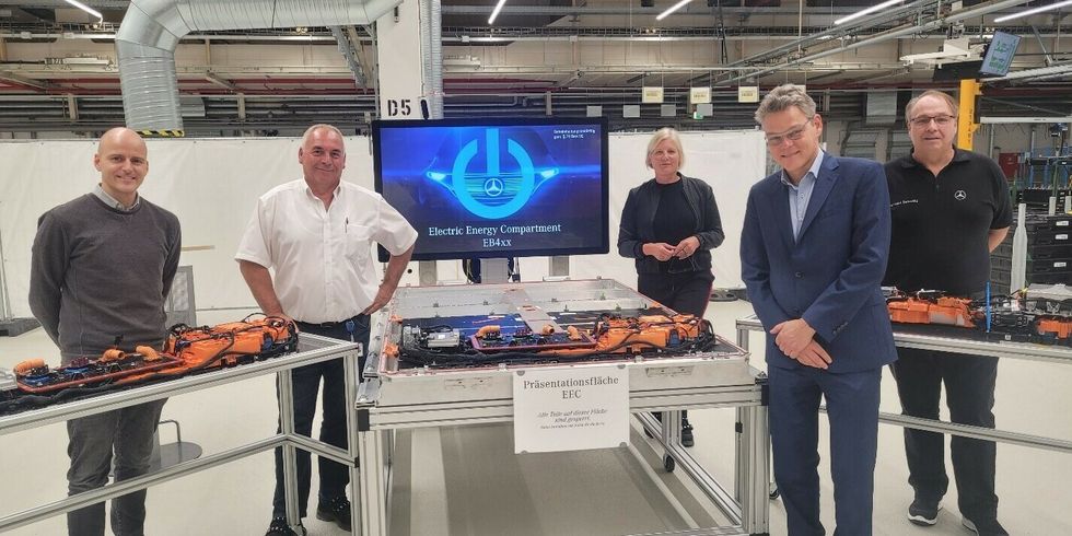 Vier Männer und eine Frau stehen in einer großen Industriehalle um Tische mit Elektroteilen.