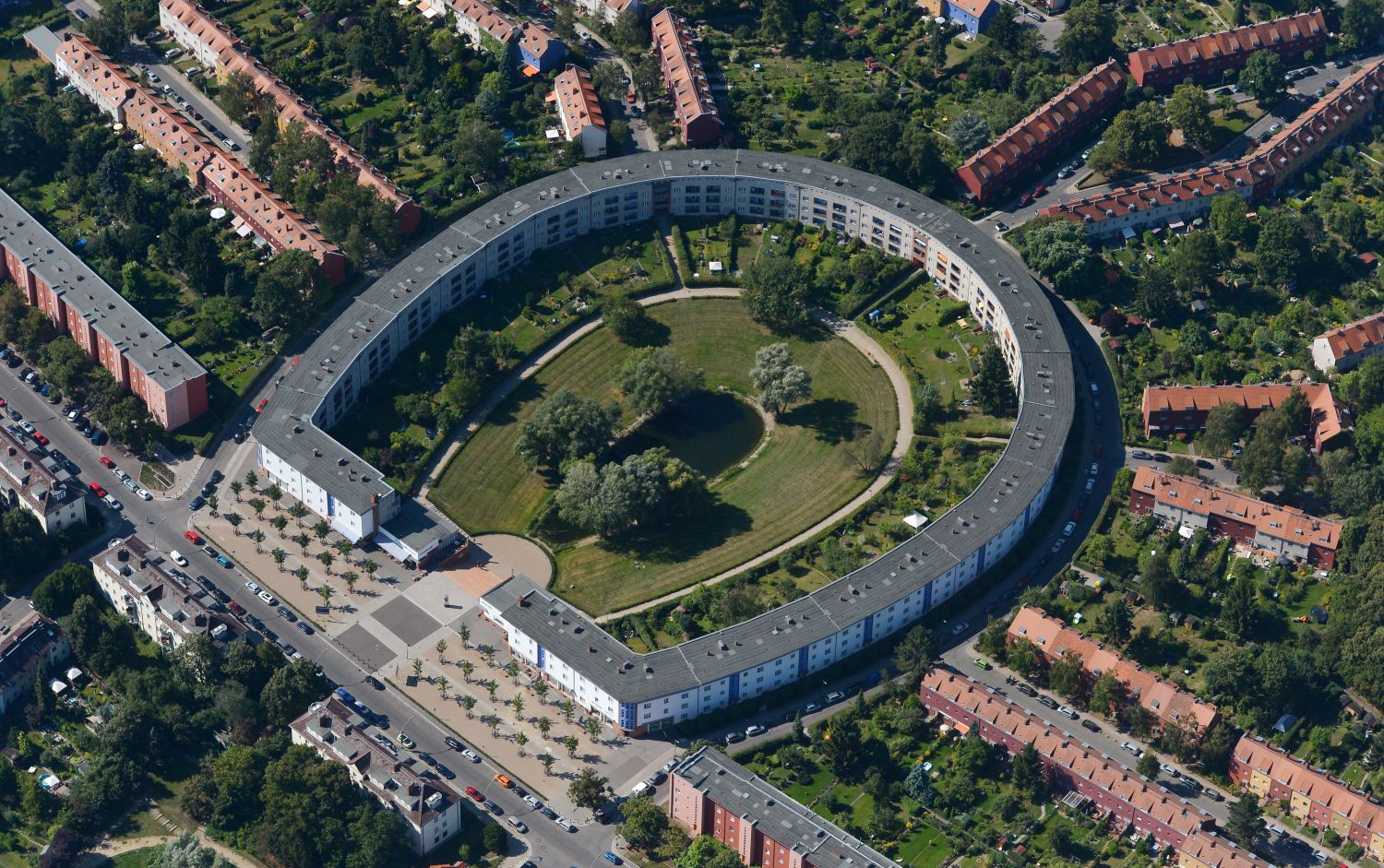 Luftbildaufnahme der Hufeisensiedlung. In der Mitte des Bildes die Hufeisensiedlung mit einem grauen Dach. Der Innenhof der Siedlung ist begrünt und es gibt einen kleinen Teich in der Mitte. Rechts und links, sowie oberhalb der Hufeisensiedlung gehen strahlenförmig weitere Reihen Häuser weg, von denen nur die roten Dächer zu sehen sind