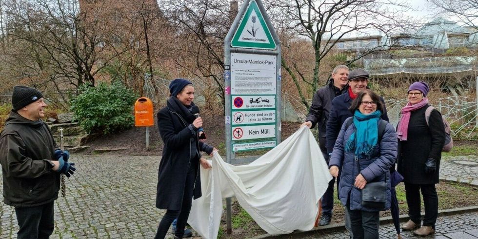 Mehrere Personen stehen an einem Parkeingangsschild. Zwei Personen nehmen eine weiße Plane von dem Schild herunter.