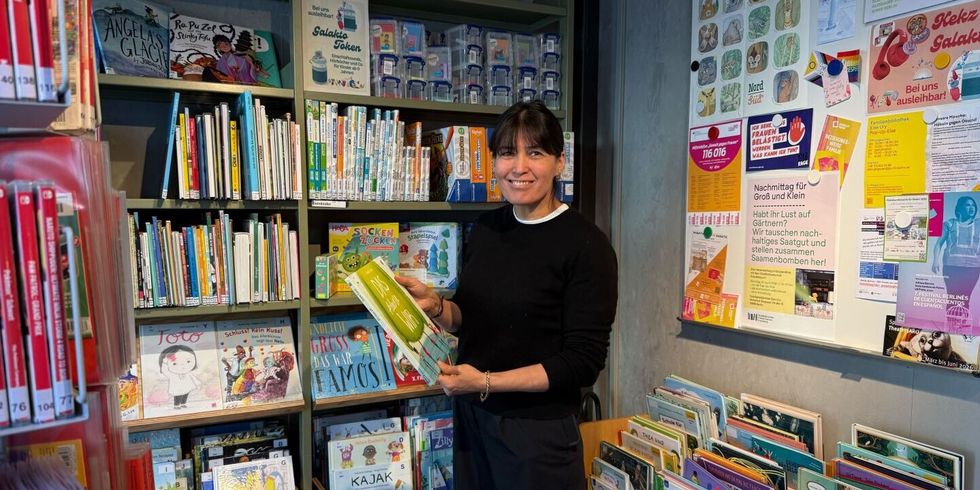 Mohidil Shamshidinova steht in der Else-Ury-Bibliothek vor einem Regal mit zahlreichen Kinderbüchern. Sie hält ein großformatiges Buch mit grünem Einband in den Händen und blickt darauf. Im Hintergrund sind weitere Bücherregale mit bunten Buchrücken zu sehen. Rechts an der Wand hängen mehrere Plakate und Informationsblätter, die farbig gestaltet sind. Links im Bild befinden sich weitere Bücherregale mit roten und weißen Buchrücken. Die Szene wirkt hell und freundlich.