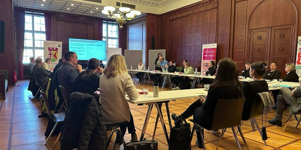 Viele Personen sitzen an Tischen. Am Ende der Tische wird eine Präsentation gezeigt.