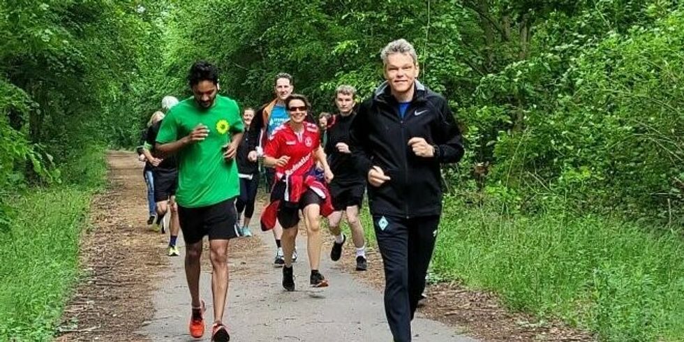 Mehrere Personen in sportlicher Bekleidung joggen auf einem Weg zwischen Bäumen.