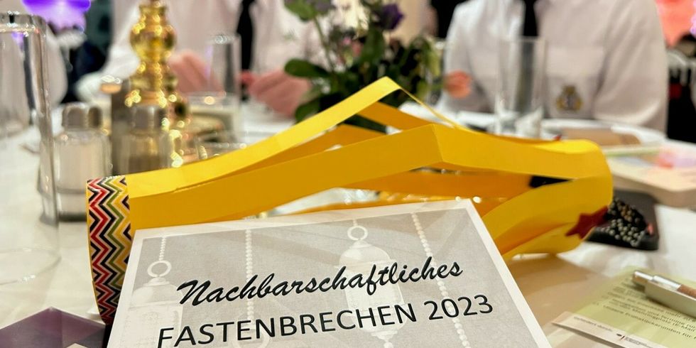 Geschmückter Tisch, im Vordergrund ein Flyer für das Nachbarschaftliche Fastenbrechen 2023, im Hintergrund zwei Gäste der Veranstaltung