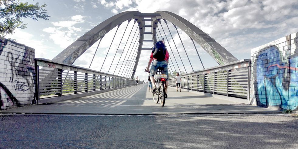 Eine fahrradfahrende Person von hinten, die über eine Zähler fährt, der bei einer Brückeneinfahrt in den Boden eingelassen ist.