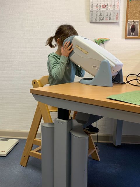Ein Kind sitzt auf einem Holzstuhl und blickt mit dem Gesicht in ein augenärztliches Untersuchungsgerät, das auf einem Tisch steht. Das Kind trägt einen langärmligen Pullover. Unter dem Tisch steht eine Personenwaage auf dem Boden.