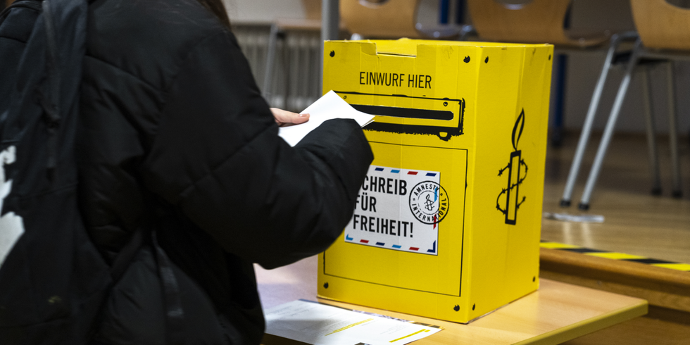 Person wirft Brief in einen als Brifkasten dekorierten Pappkarton mit der Aufschrift "Einwurf hier - Schreib für Freiheit"