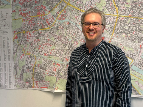Aaron Schaar steht mit neutralem Gesichtsausdruck und gestreiftem Hemd vor einer großformatigen Stadtkarte an der Wand.