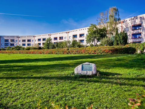 Im Vordergrund eine hellgrüne Wiese mit einem hellgrauen Stein auf dem eine grüne Plakette ist. Dahinter im Halbrund eine Häuserfront. Die Hauswände sind weiß. die Balkone sind nach innen gebaut, mit jeweils blauen Rückwänden