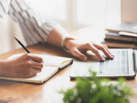 Nahaufnahme der Hände einer Frau, die am Laptop arbeitet und Notizen mit einem Stift macht