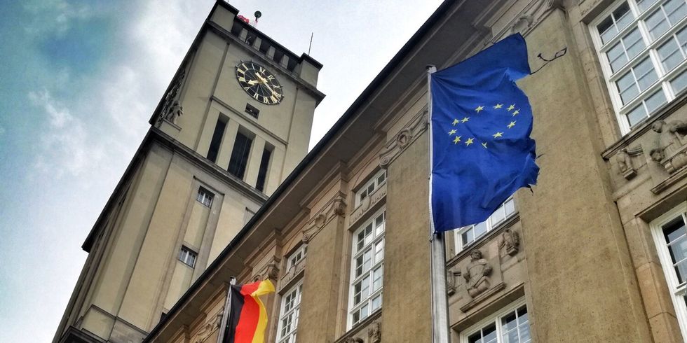 Vor einem großen Gebäude mit Uhrenturm sind Fahnenmäste mit Flaggen. Im Vordergrund ist eine blaue Flagge mit gelben Sternen in einer Kreisformation zu sehen.