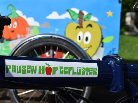 Ein Smoothiebike mit der Aufschrift Pausenhofgeflüster, im Hintergrund ein Comic mit Obst-Figuren