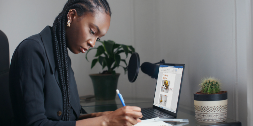 Eine Frau arbeitet zu Hause an ihrem Schreibtisch bei aufgeklapptem Notebook und macht sich Notizen in ihrem Notizbuch.