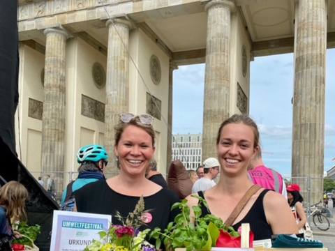 Zwei Frauen stehen vor dem Brandenburger Tor, sie halten Blumen und eine Urkunde des Umweltfestivals