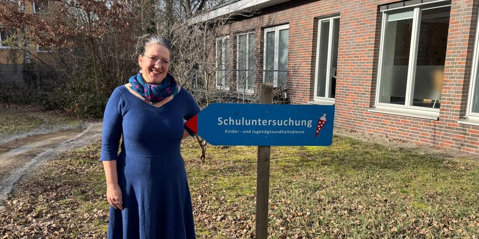 Eine Person mit blauem Kleid, gemustertem Schal und dunklen Stiefeln steht auf Laub und Gras neben einem Holzpfosten mit einem blauen Richtungsschild. Das Schild trägt den Schriftzug 'Schuluntersuchung Kinder- und Jugendgesundheitsdienst'. Im Hintergrund ist ein Backsteingebäude mit großen Fenstern und kahlen Bäumen zu sehen.