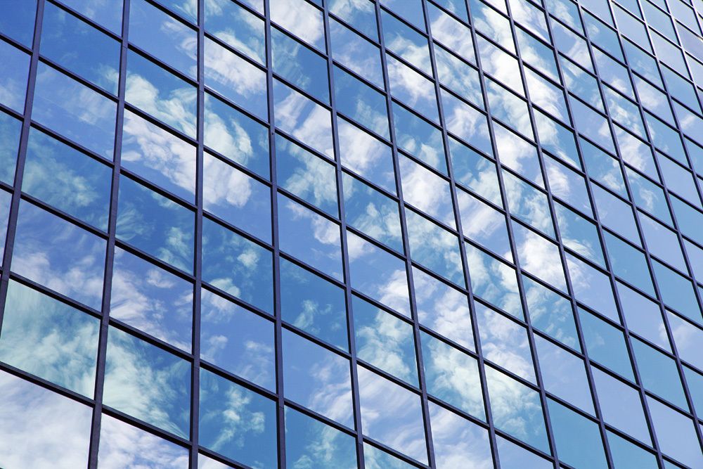 Ein Blick von schräg unten auf die riesige Glasfassade eines Gebäudes, die das gesamte Foto ausfüllt. Schmale schwarze Rahmen unterteilen die Fläche in gleichgroße Quadrate. Der blaue Himmel mit weißen Wolken spiegelt sich darin. Die Scheiben jeder zweiten waagerechten Reihe erscheinen blau-grünlich getönt.