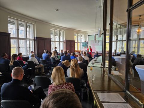 In einem Raum sitzen zahlreiche Personen, eine Frau hält einen Vortrag 