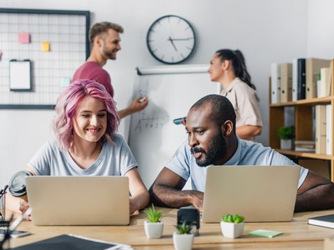 Zwei Personen sitzen im Büro vor ihren Laptops, im Hintergrund stehen zwei Menschen vor einem Flipchart