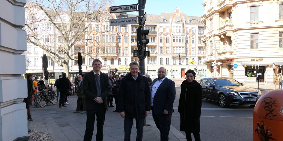 Vier Personen stehen nebeneinander vor einem Mast mit Straßenschildern. Das Schild Kaiser-Wilhelm-Platz ist rot durchgestrichen und darunter ist ein neues Schild Richard-von-Weizsäcker-Platz.