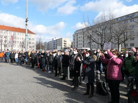 Viele Menschen stehen versammelt draußen auf einem Platz und schauen alle zusammen in eine Richtung.