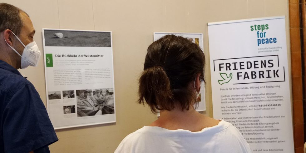 Eine Frau und ein Mann stehen der Kamera abgewendet und schauen auf einen Aufsteller und aufgehängte Bilder mit Texten. Auf dem Aufsteller steht "Steps for Peace" und "Friedensfabrik".