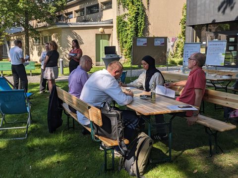 Personen arbeiten an Holztischen im Freien mit Flipcharts im Hintergrund