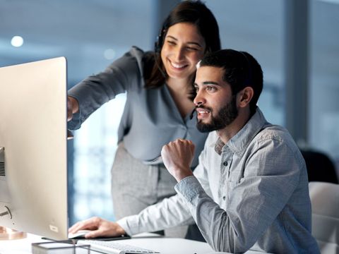 Ein Mann sitzt an einem PC-Arbeitsplatz und eine Frau steht neben ihm und zeigt ihm etwas auf dem Bildschirm