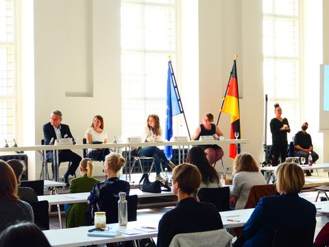 Drei Frauen und ein Mann am Tisch, im Vordergrund Zuhörer