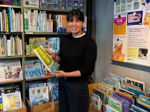 Mohidil Shamshidinova steht in der Else-Ury-Bibliothek vor einem Regal mit zahlreichen Kinderbüchern. Sie hält ein großformatiges Buch mit grünem Einband in den Händen und blickt darauf. Im Hintergrund sind weitere Bücherregale mit bunten Buchrücken zu sehen. Rechts an der Wand hängen mehrere Plakate und Informationsblätter, die farbig gestaltet sind. Links im Bild befinden sich weitere Bücherregale mit roten und weißen Buchrücken. Die Szene wirkt hell und freundlich.