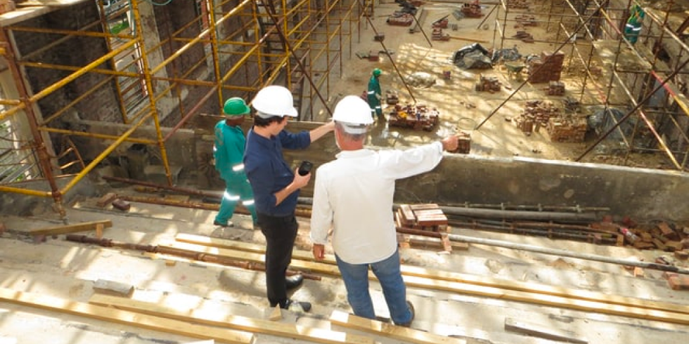 Zwei Arbeiter befinden sich auf einer Baustelle, stehen auf einem Gerüst und beraten sich.
