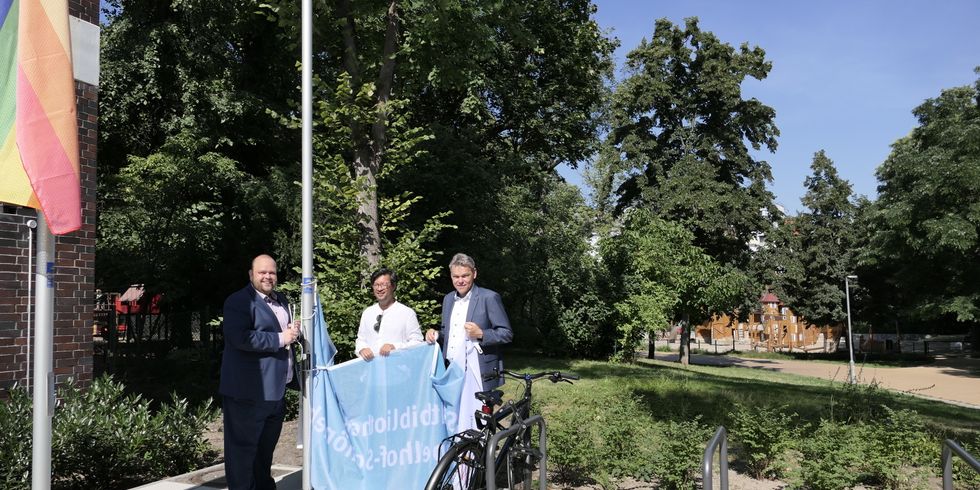 Drei Männer halten eine Flagge an einem Fahnenmast. Links davon ist ein weiterer Fahnenmast mit einer Regenbogenflagge. Im Hintergund ist ein Spielpltz.