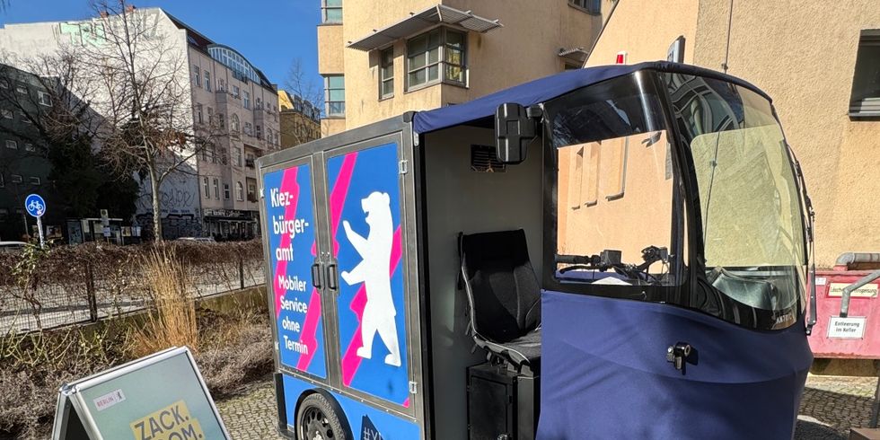 Lastenfahrrad mit Kofferaufbau mit blauem Werbeaufdruck und offenem Fahrersitz steht auf gepflastertem Gehweg neben einem aufgestellten Werbeschild