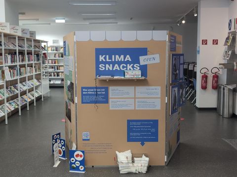 Eine Ausstellungswand mit der großen Aufschrift "Klimasnack", Im Hintergrund eine Bibliothek