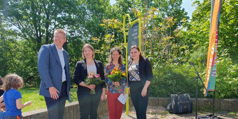 3 Frauen und ein Mann stehen vor einer Informationstafel auf einem Platz, im Hintergrund Bäume