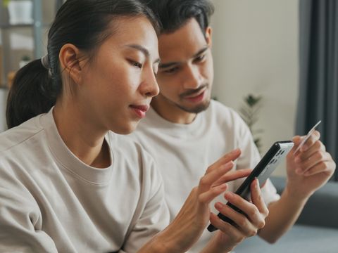 Happy young Asian couple sit on couch spend time together have fun use smartphone devices shopping use credit card online payment at home. Young husband and lifestyle domestic activity concept.