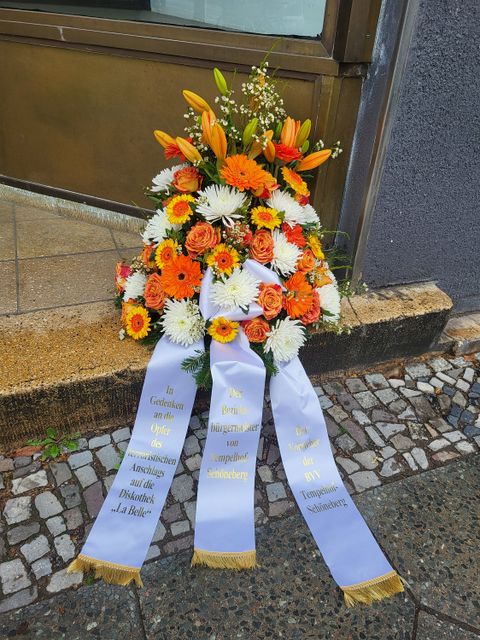 Blumen mit einem Band wo drauf steht "In Gedenken an die Opfer des terroristischen Anschlags auf die Diskothek La Belle"
