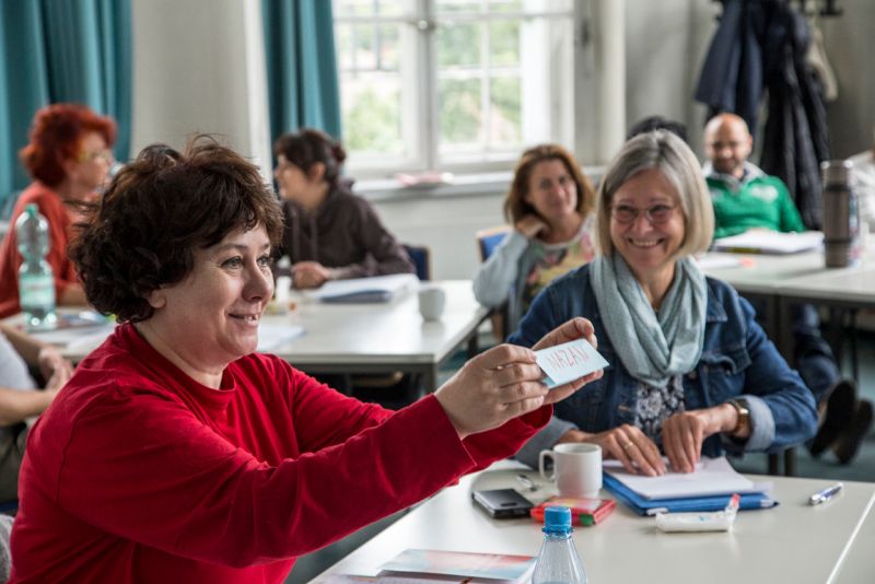 Zwei Frauen sitzen vorne an einem Tisch, dahinter drei Frauen und ein Mann an weiteren Tischen.