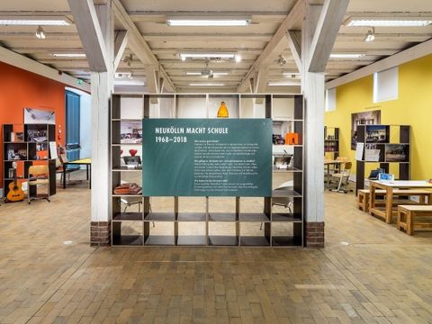 Blick in die Ausstellung Neukölln macht Schule, es ist die Eingangswand mit enleitendem Text zu sehen, sowie verschiedene Schultische und Schulbänke.