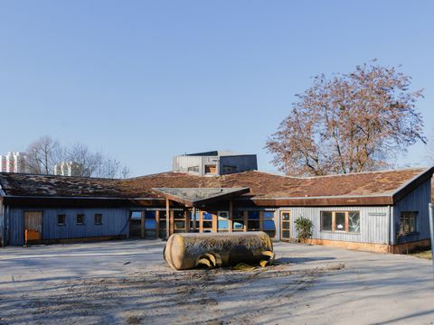 Über Eck gebauter Flachbau in Holzbauweise mit Gründach. Im Hof ein großer Kessel
